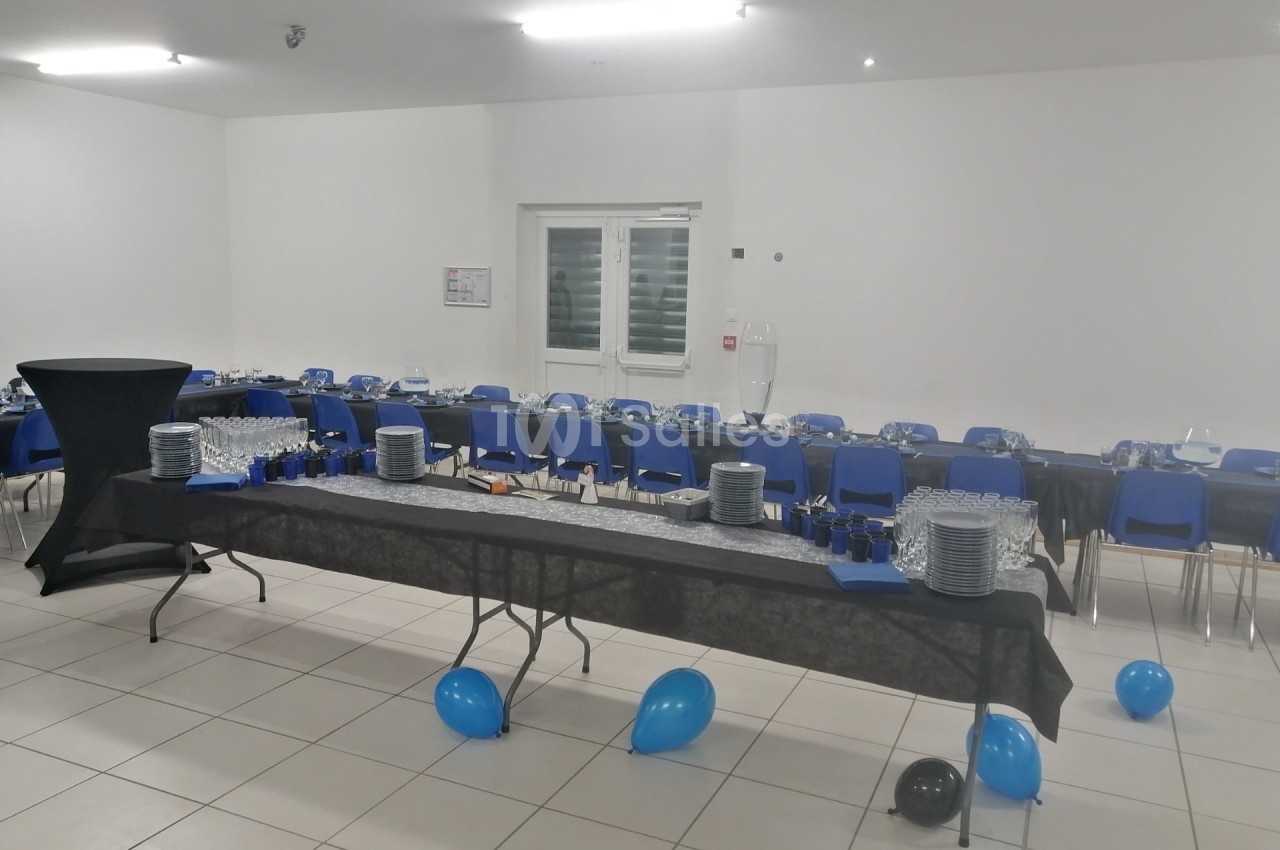 Salle décorée pour un événement avec tables alignées, nappes noires, vaisselle disposée et ballons bleus et noirs au sol.