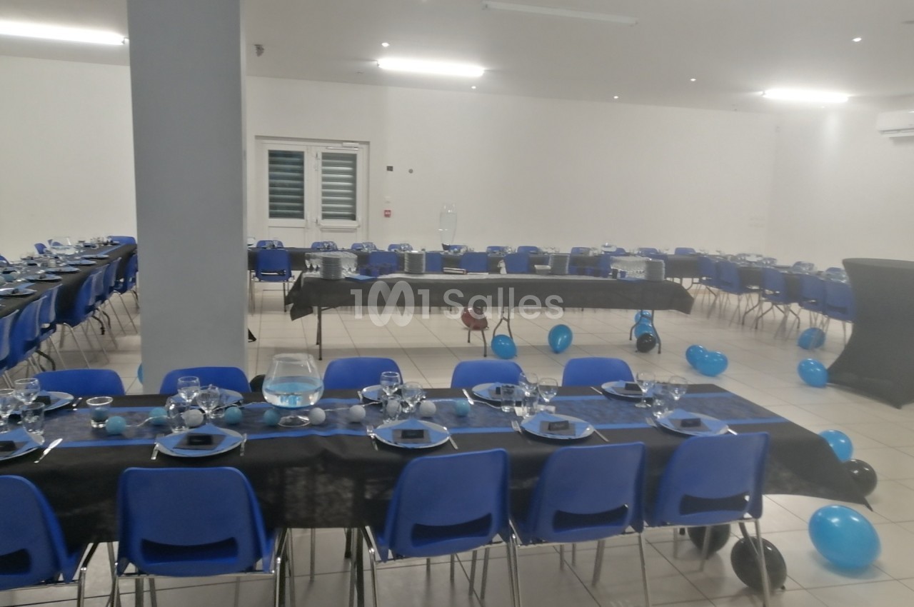 Salle décorée pour un événement avec des tables alignées, nappes noires, vaisselle, et ballons bleus au sol.