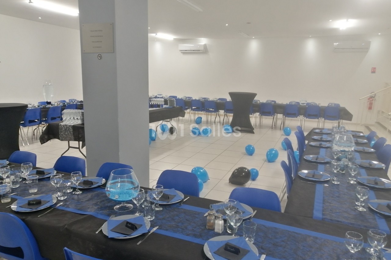 Salle de réception décorée avec des tables dressées en noir et bleu, des ballons au sol et des chaises assorties.
