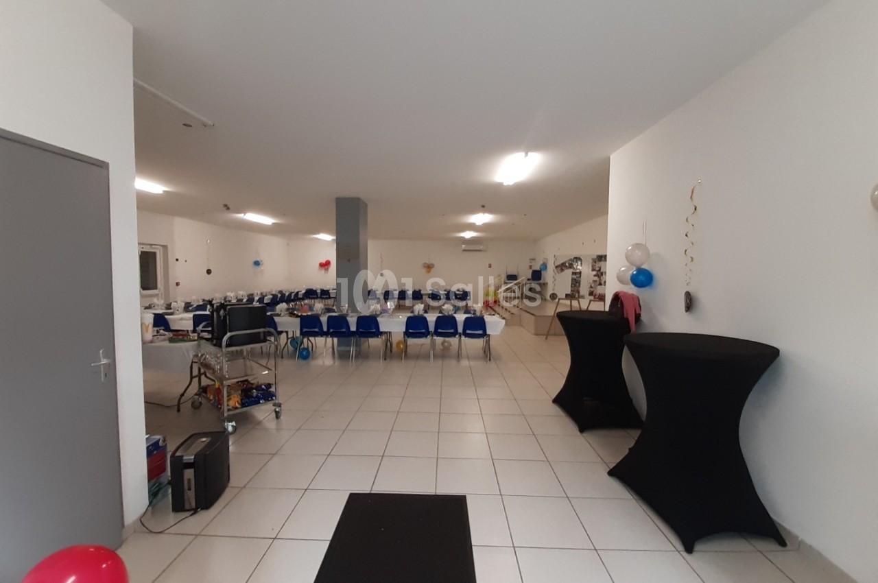 Salle spacieuse avec tables et chaises alignées, décorée de ballons colorés, prête pour un événement.