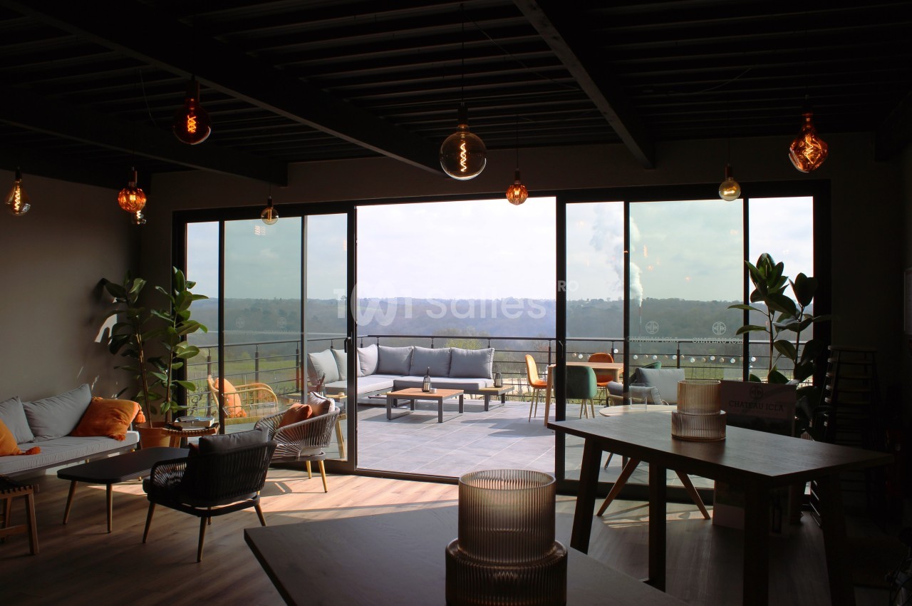 Salle lumineuse avec grandes baies vitrées donnant sur une terrasse aménagée et vue sur un paysage vallonné.