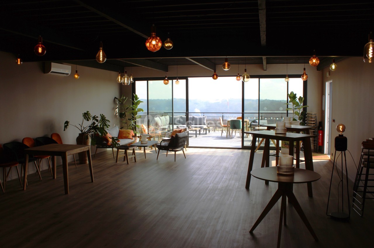 Salle lumineuse avec grandes baies vitrées, mobilier en bois, plantes et vue sur une terrasse extérieure.