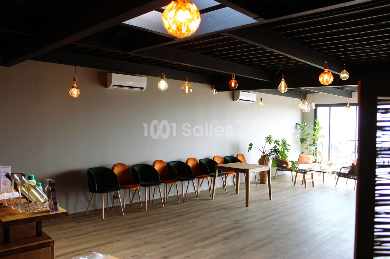 Salle lumineuse avec rangée de chaises, plantes, table en bois et suspensions décoratives sous un plafond sombre.