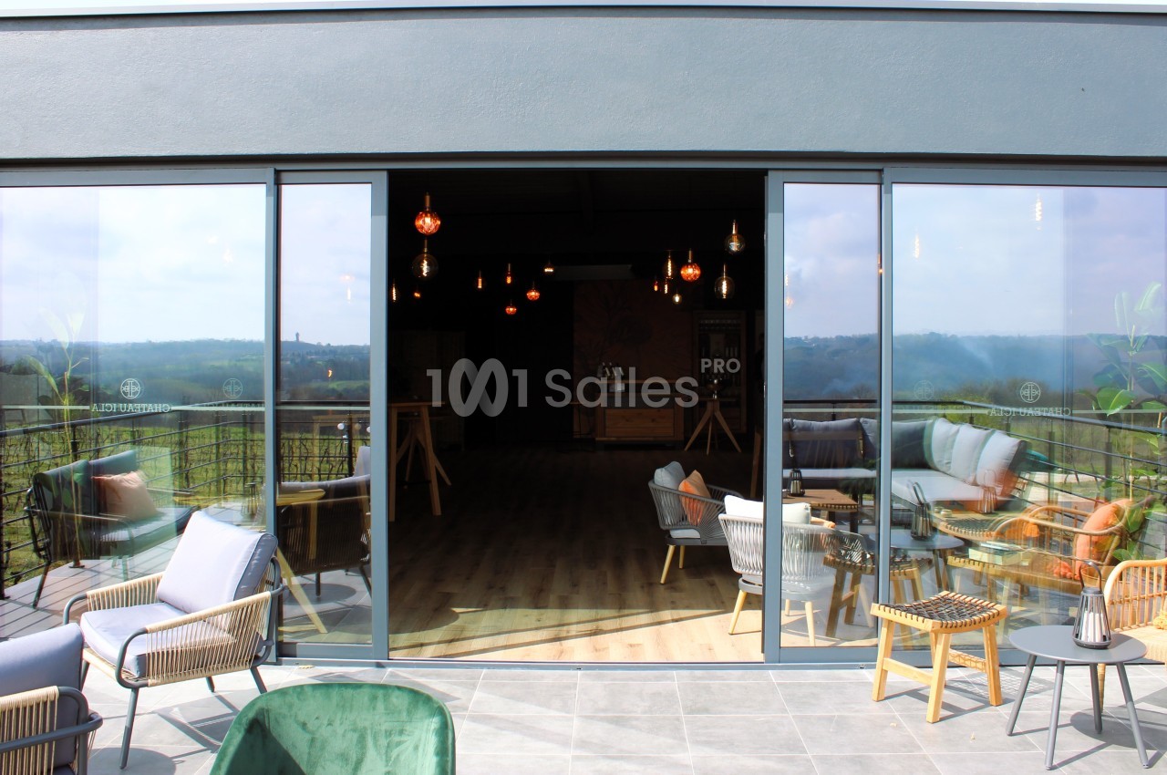 Vue d'une terrasse aménagée avec des fauteuils et une table, donnant sur une salle intérieure lumineuse et un paysage…