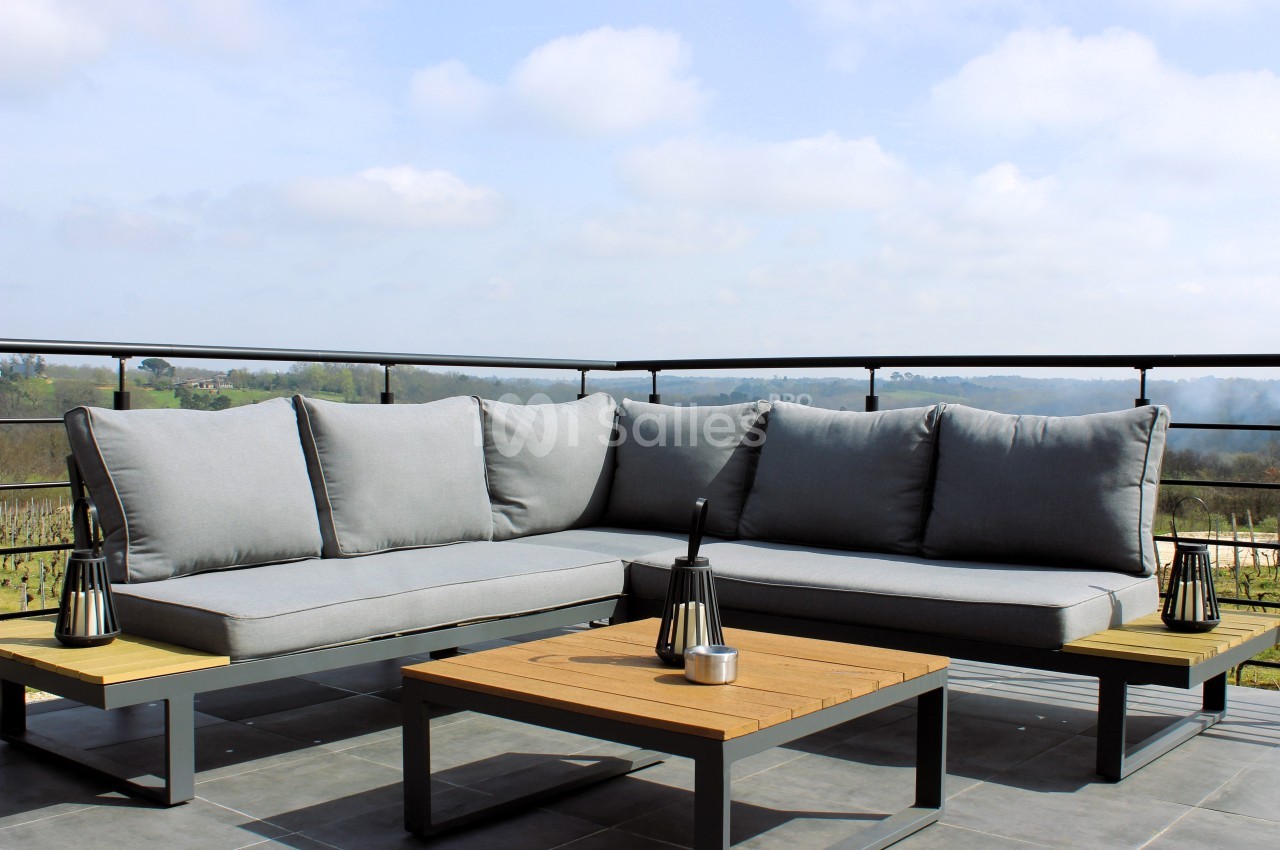 Canapé d'angle gris avec table basse en bois sur une terrasse offrant une vue dégagée sur la campagne.