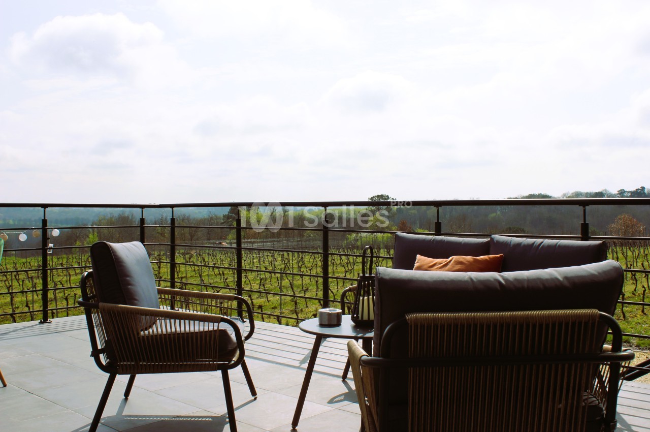 Terrasse avec mobilier en rotin, table basse et vue dégagée sur un paysage de vignobles sous un ciel légèrement nuageux.