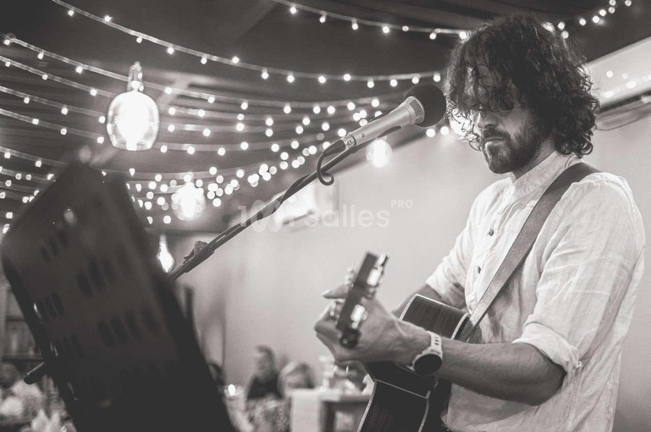 Un musicien joue de la guitare et chante dans un espace décoré de guirlandes lumineuses, devant un public assis.