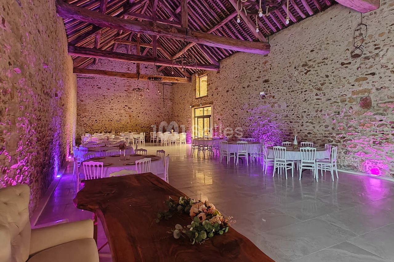 Salle en pierre avec poutres apparentes, tables et chaises blanches, éclairage violet et décoration florale sur une table en…