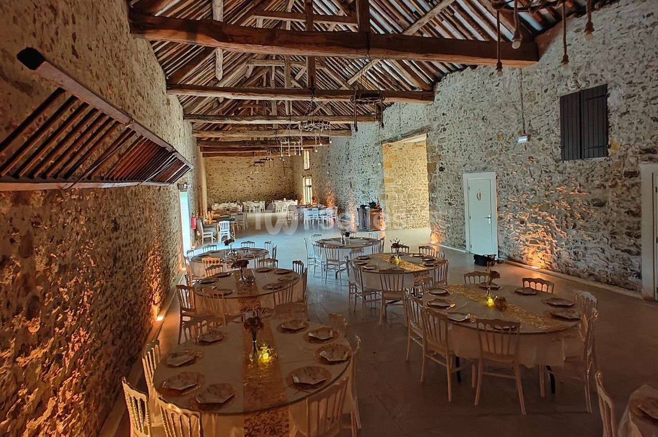 Salle en pierre avec poutres apparentes, tables rondes dressées pour un événement, éclairage chaleureux.