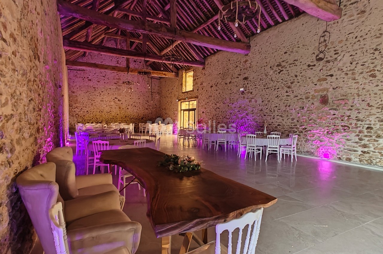 Salle en pierre avec poutres apparentes, table en bois massif, chaises blanches et éclairage violet tamisé.
