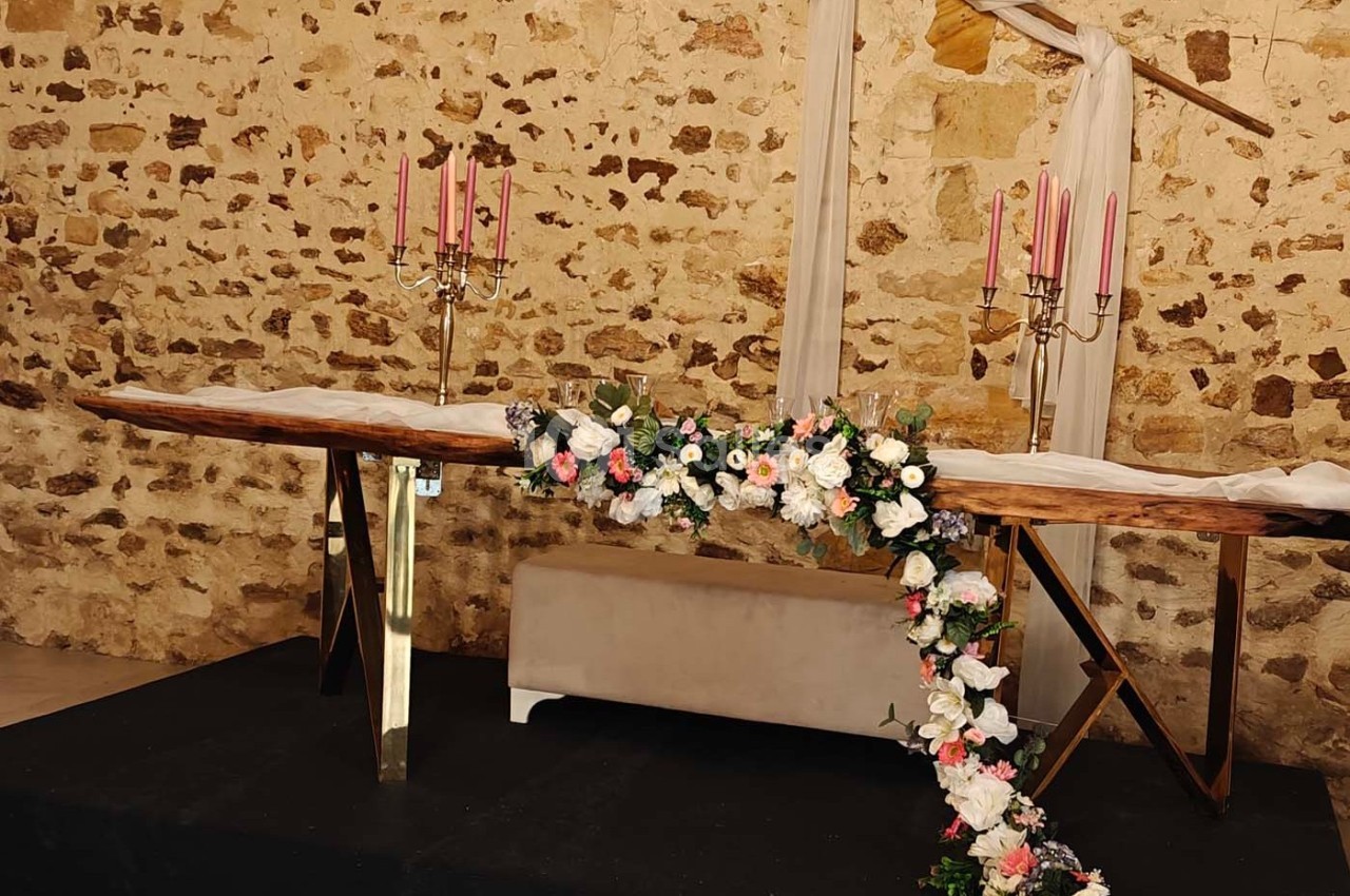 Table en bois décorée de chandeliers roses et de guirlandes florales, placée devant un mur en pierre apparente.