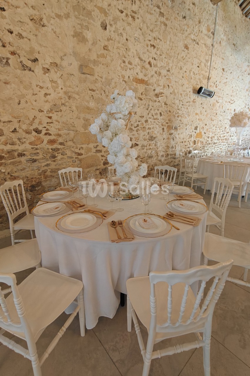 Table ronde élégamment dressée avec nappes blanches, vaisselle raffinée et centre de table floral dans une salle en pierre.