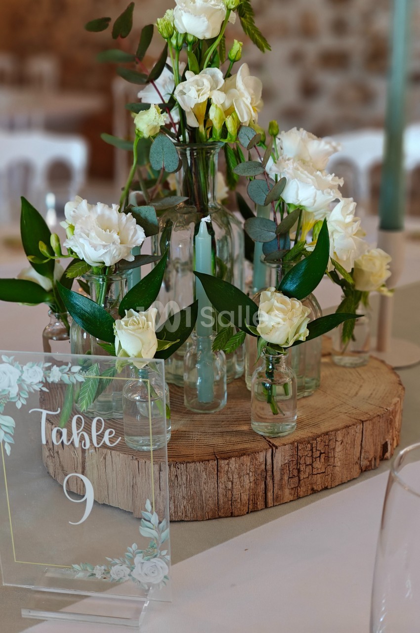 Centre de table avec des fleurs blanches et feuillages dans des vases en verre, posé sur une rondelle de bois.