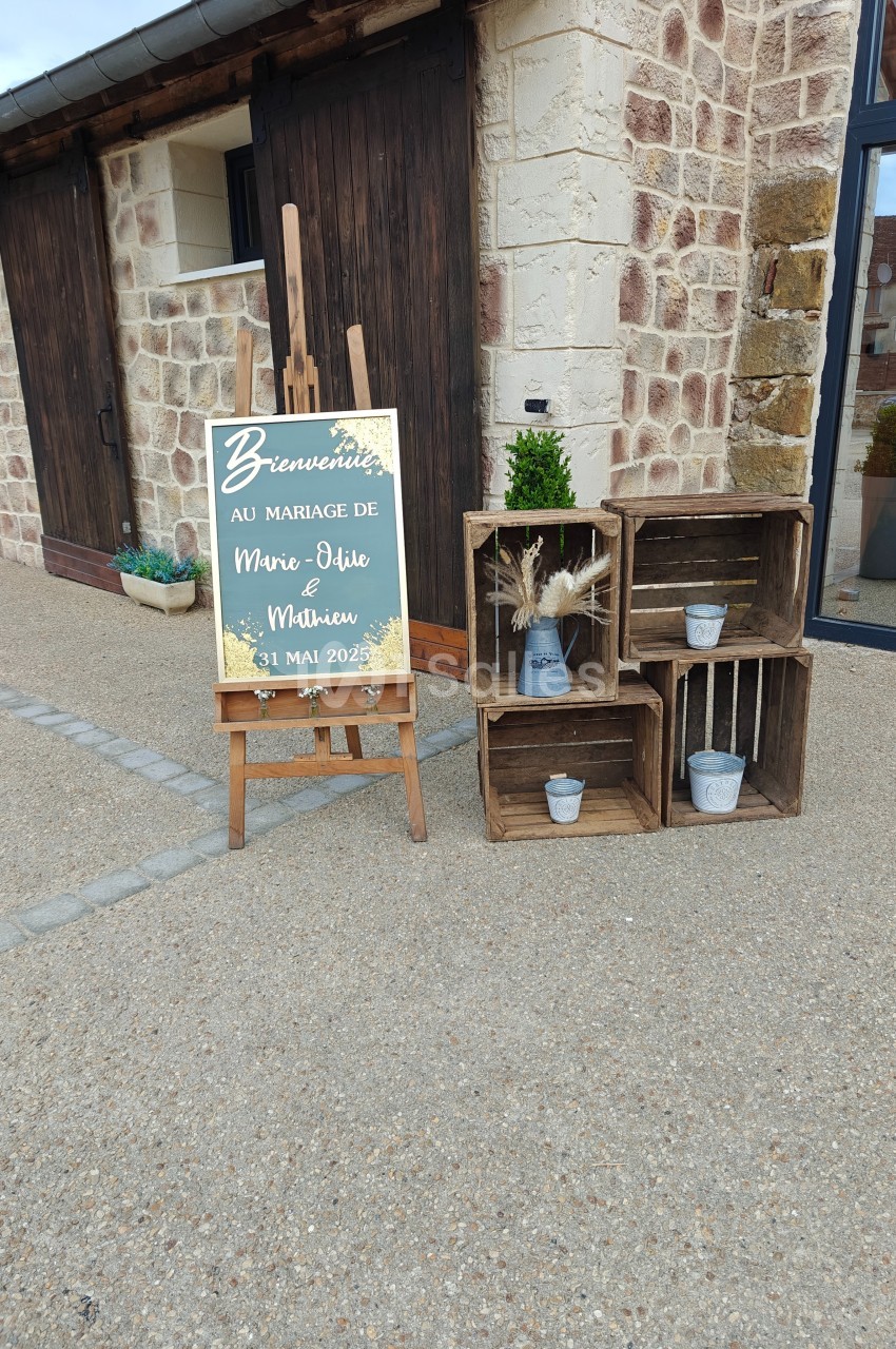 Panneau de bienvenue pour un mariage, posé sur un chevalet près de caisses en bois décoratives devant un bâtiment en pierre.