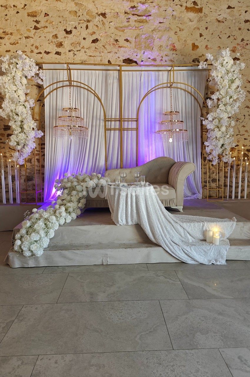 Décor de mariage avec canapé beige, rideaux blancs, arches dorées, fleurs blanches et chandeliers suspendus.