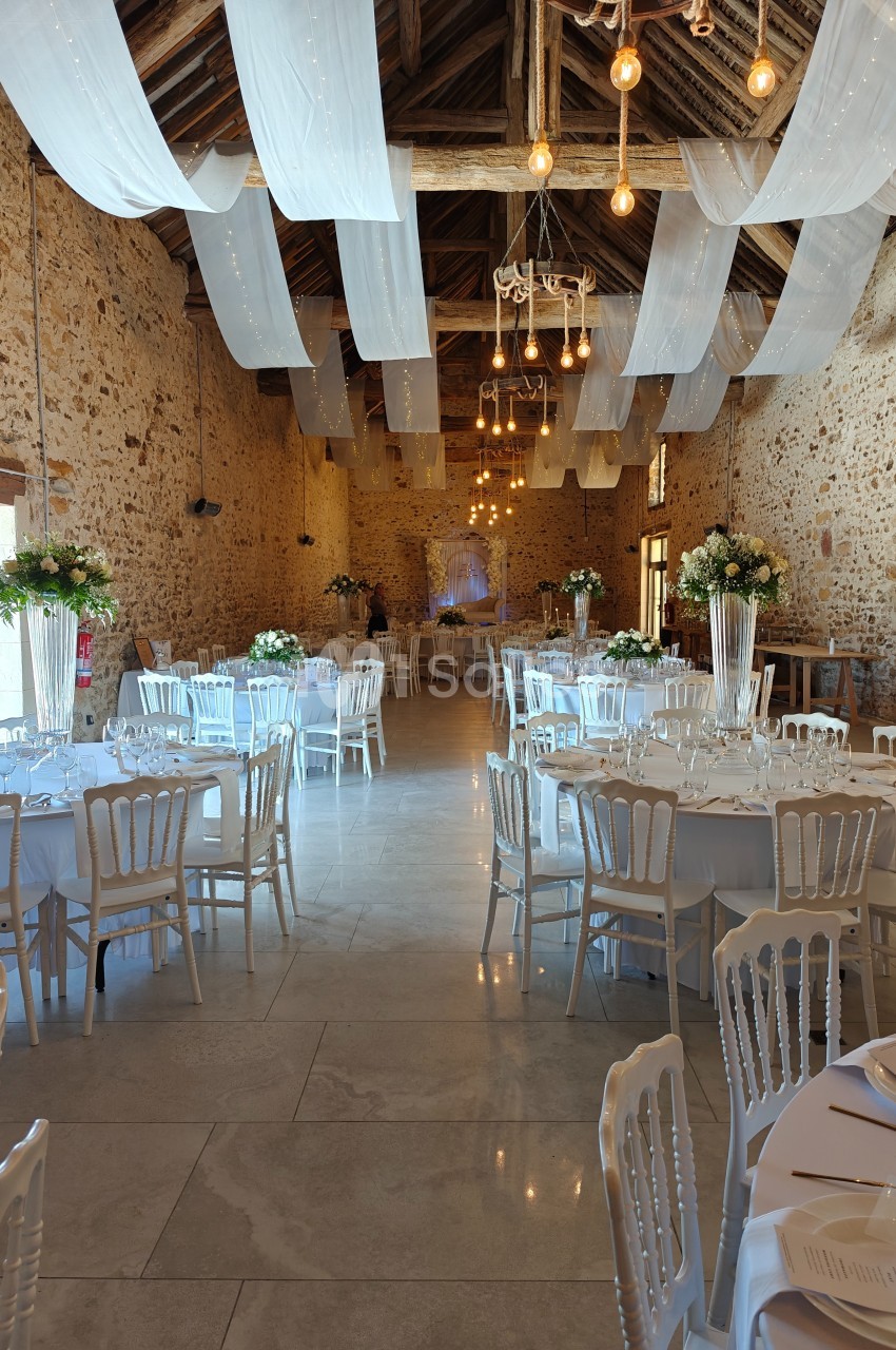 Salle de réception en pierre avec tables rondes décorées, chaises blanches et plafonds ornés de voilages et lumières…