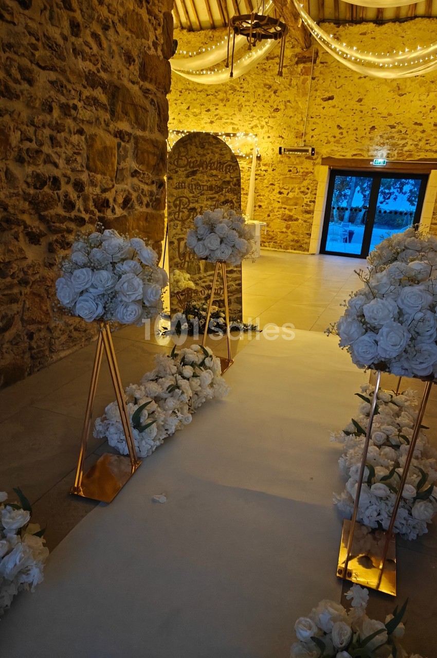 Allée décorée de fleurs blanches dans une salle en pierre éclairée par des guirlandes lumineuses.