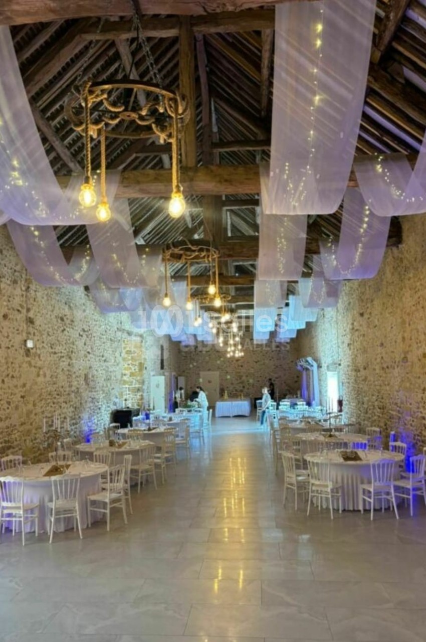 Salle en pierre avec plafond en poutres apparentes, décorée de voilages et guirlandes lumineuses, tables rondes dressées.