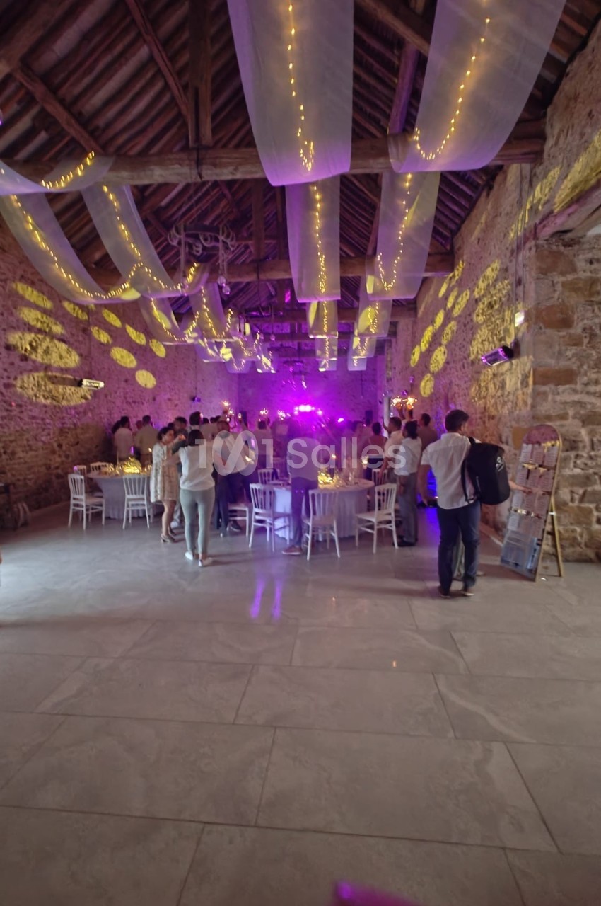 Salle en pierre avec des guirlandes lumineuses suspendues, des invités debout et assis autour de tables éclairées.