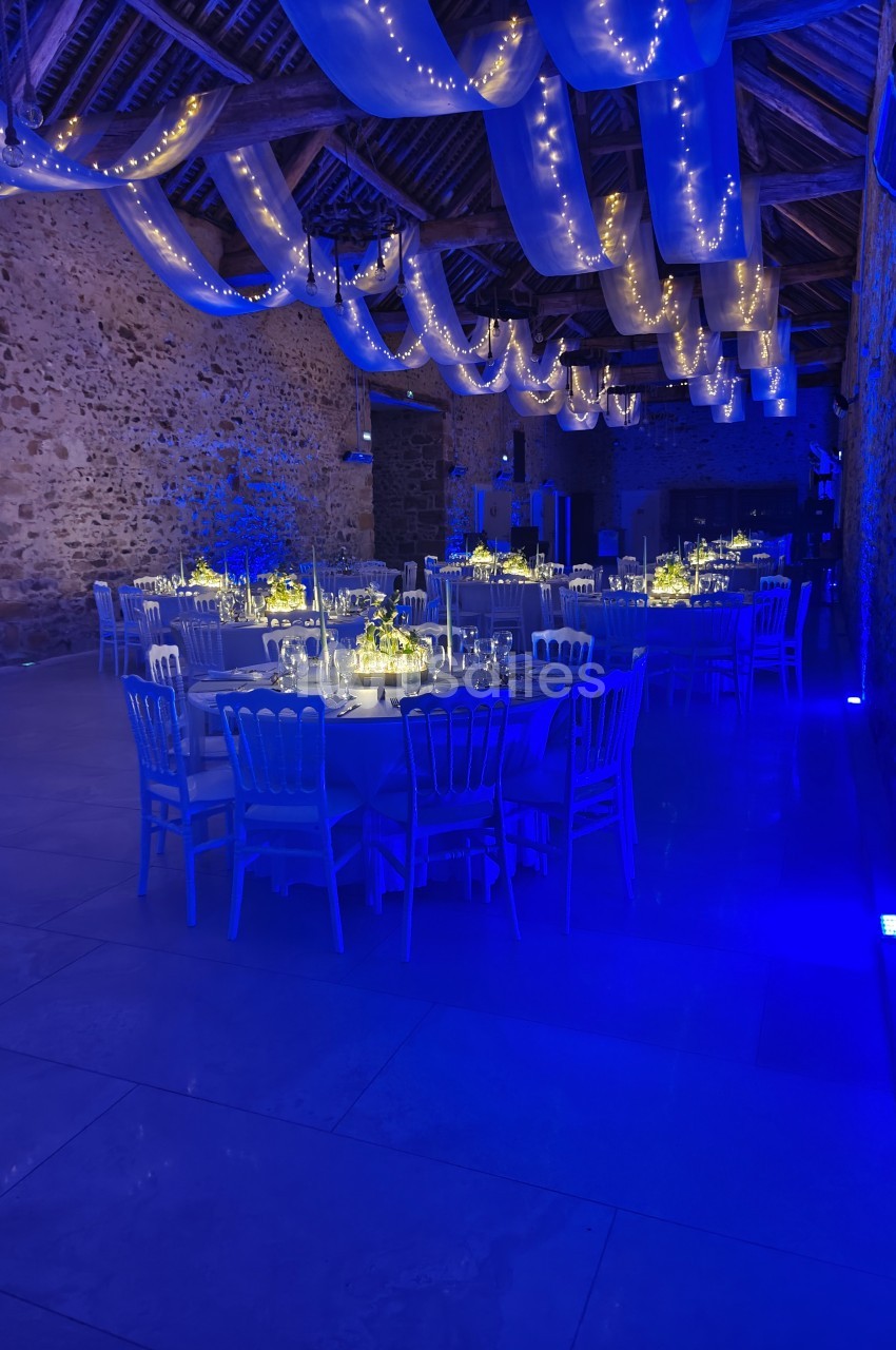 Salle de réception en pierre éclairée en bleu, décorée de guirlandes lumineuses et tables rondes dressées.