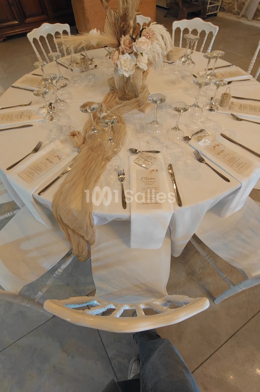 Table ronde élégamment dressée avec nappes blanches, menus, verres, couverts et décoration florale beige et blanche.