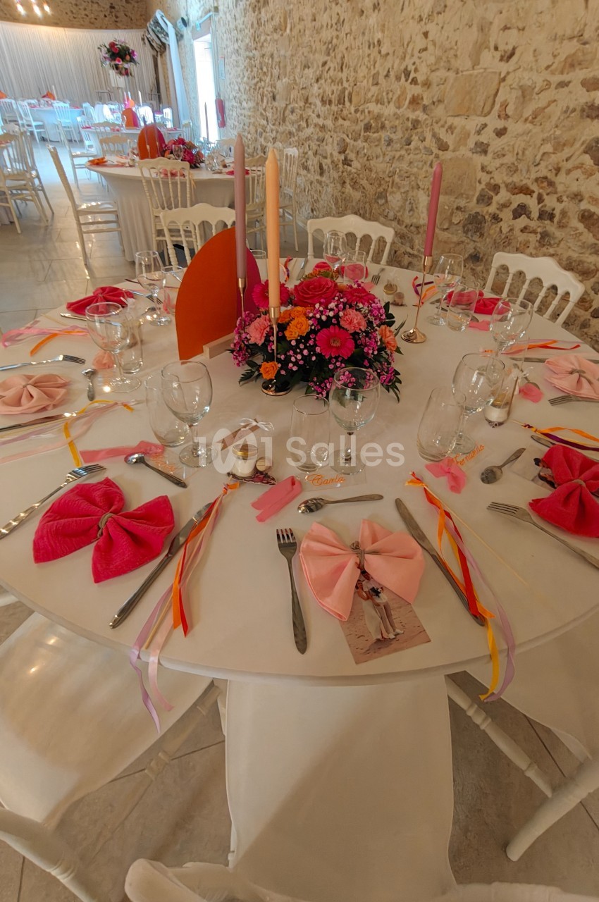 Table ronde décorée pour un événement, avec nappes blanches, serviettes roses, bougies et centre de table floral coloré.