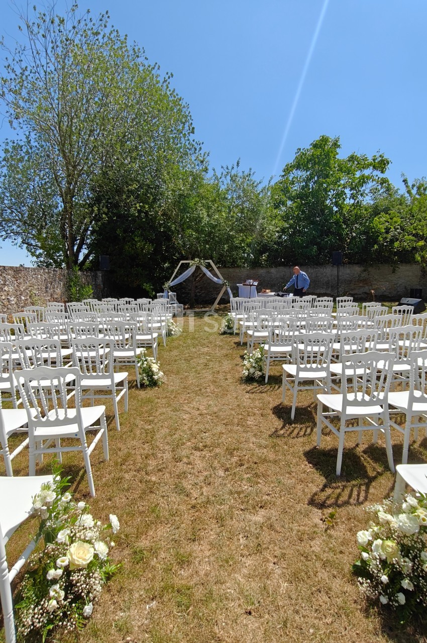 Allée centrale bordée de chaises blanches et de fleurs menant à une arche décorée pour une cérémonie en plein air.