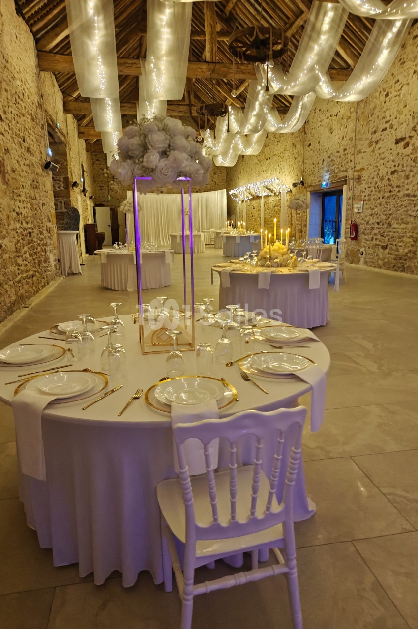 Salle de réception décorée avec des tables rondes dressées, des guirlandes lumineuses suspendues et des murs en pierre…