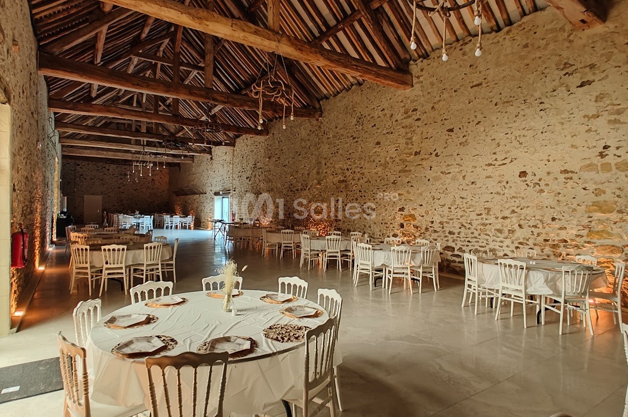 Salle de réception en pierre avec plafond en poutres apparentes, tables rondes dressées et chaises blanches.
