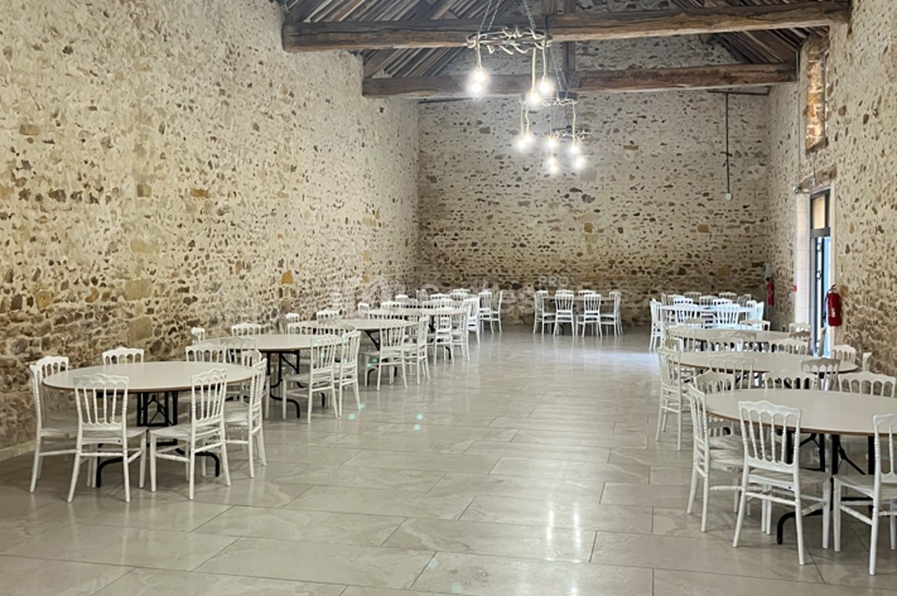 Salle en pierre avec plafond en bois, tables rondes blanches et chaises assorties, éclairée par des suspensions modernes.