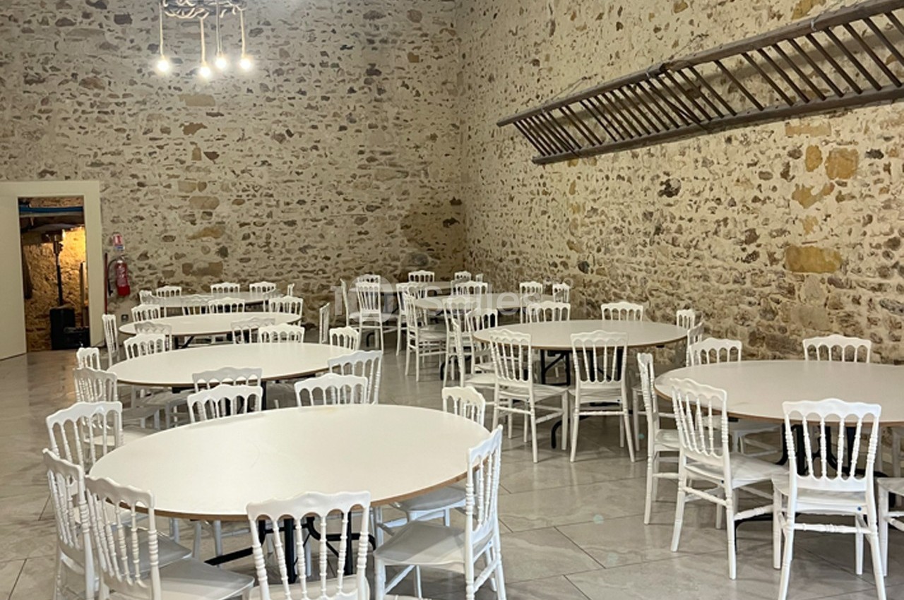 Salle en pierre avec tables rondes blanches et chaises assorties, éclairée par des suspensions modernes.