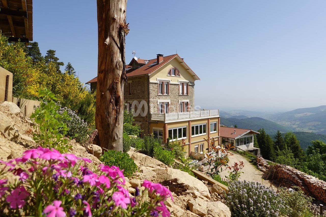 Maison en pierre entourée de végétation, avec vue sur une vallée et des collines sous un ciel dégagé.