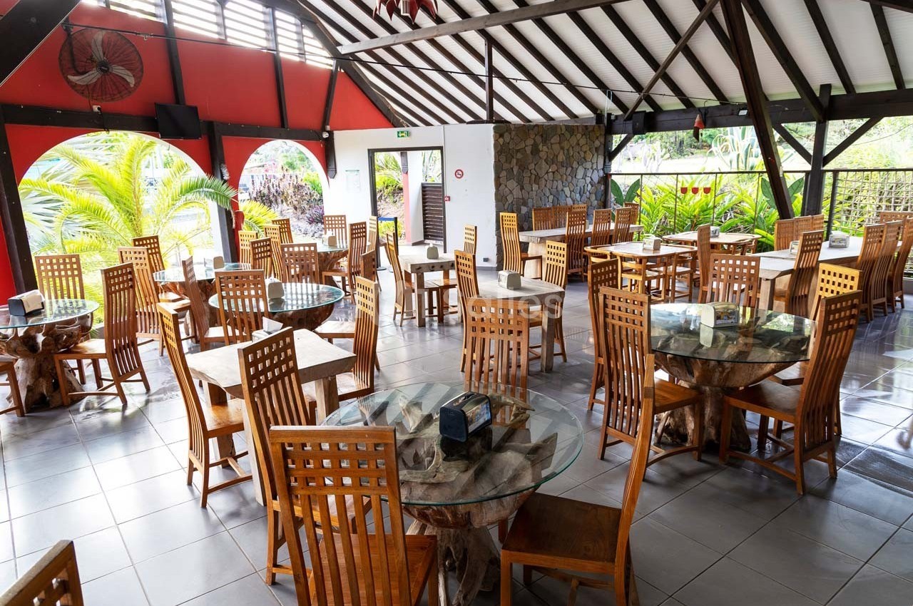 Salle de restaurant lumineuse avec des tables en verre, des chaises en bois et une vue sur un jardin tropical.