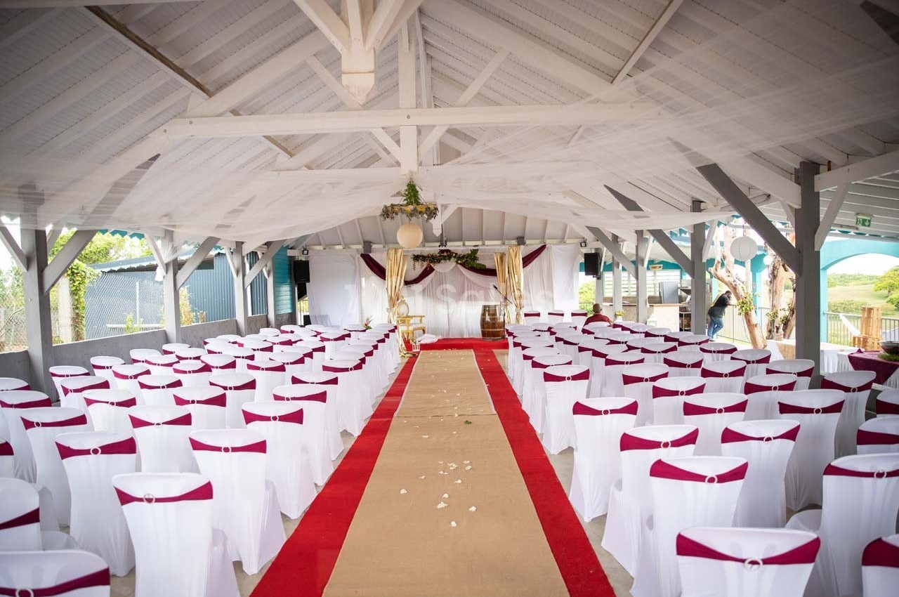 Salle décorée pour un événement avec des chaises blanches ornées de nœuds bordeaux et une allée centrale rouge.