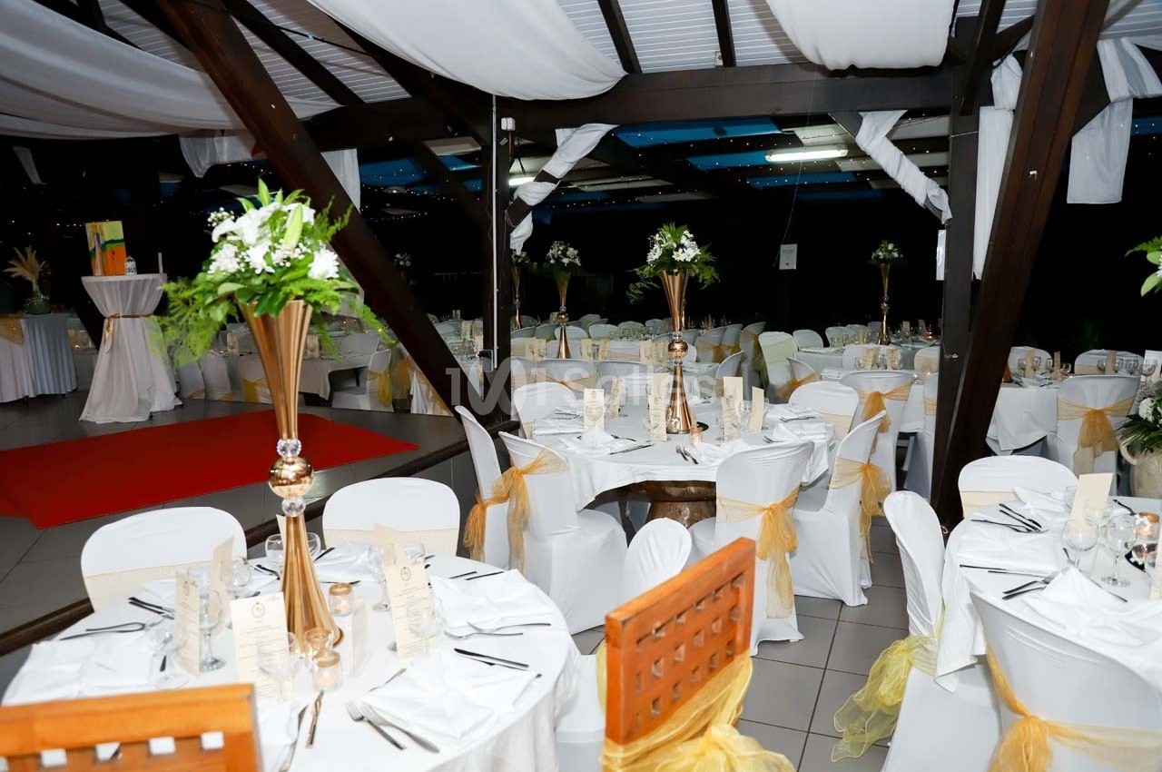 Salle de réception décorée avec des tables rondes, nappes blanches, chaises ornées de rubans jaunes et centres de table…
