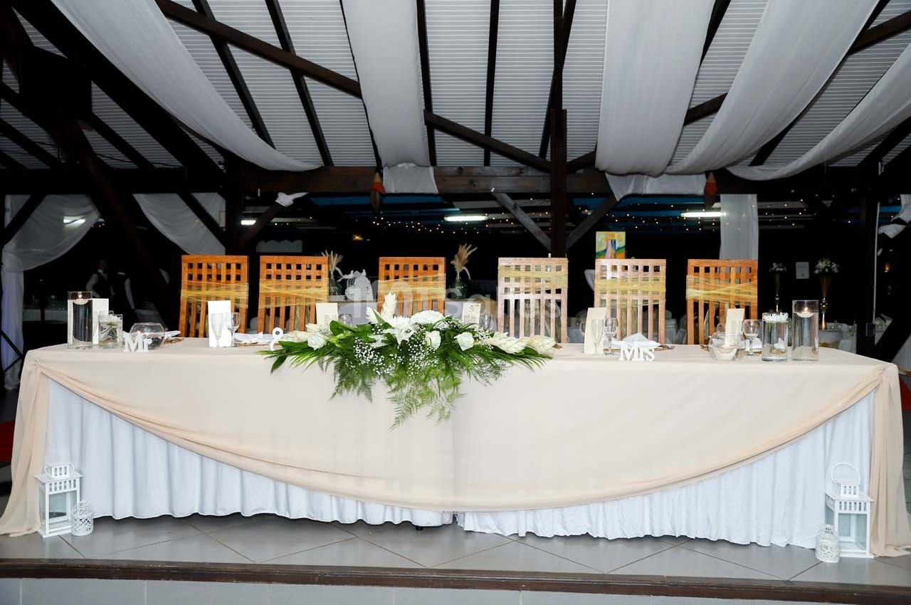 Grande table de réception décorée de nappes blanches, de lanternes, de fleurs et de chaises en bois sous un toit avec drapés.