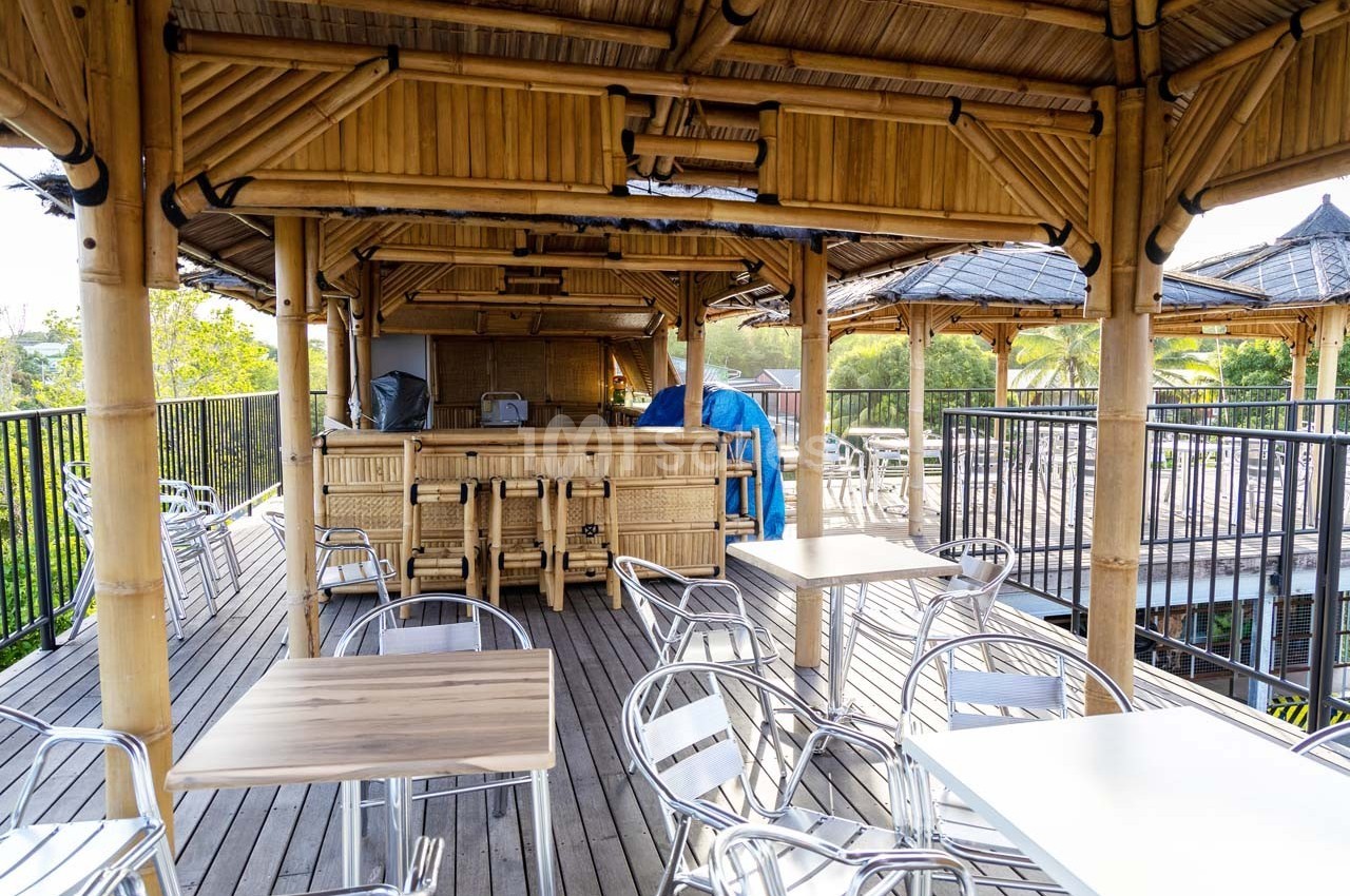 Terrasse en bois avec tables, chaises métalliques et bar en bambou, entourée de verdure et de structures en toit de chaume.