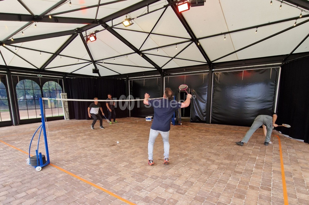 Quatre personnes jouent au badminton dans une structure couverte avec un sol en pavés et un filet central.