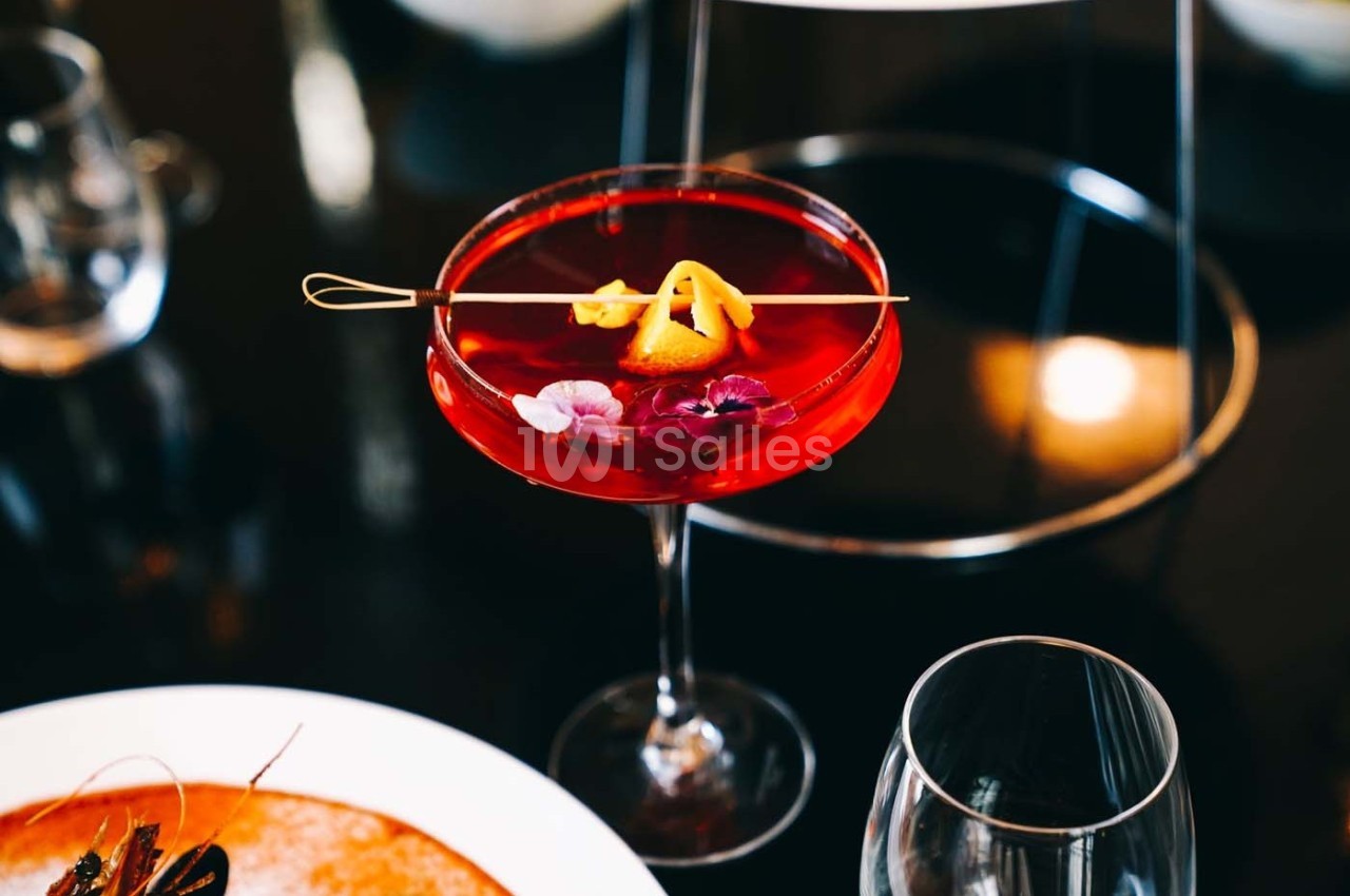 Verre à cocktail rouge décoré de fleurs comestibles et d'un zeste d'agrume, posé sur une table sombre.
