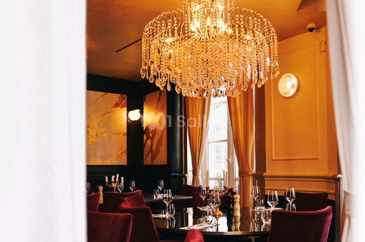 Salle de restaurant élégante avec un lustre en cristal, des rideaux beiges et des chaises rouges autour des tables.