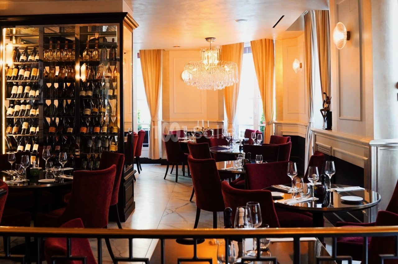 Salle de restaurant élégante avec chaises rouges, tables dressées et une cave à vin vitrée en arrière-plan.