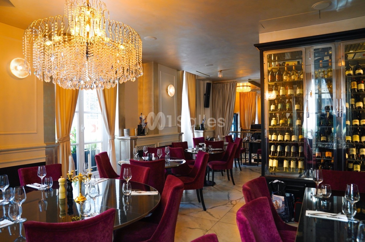 Salle de restaurant élégante avec lustre en cristal, chaises rouges, tables dressées et une cave à vin vitrée.