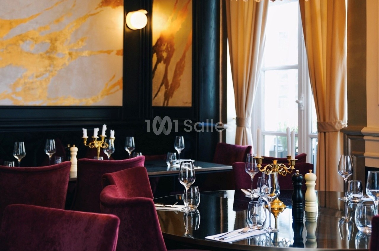 Salle de restaurant élégante avec chaises en velours rouge, tables dressées et lumière naturelle entrant par une fenêtre.