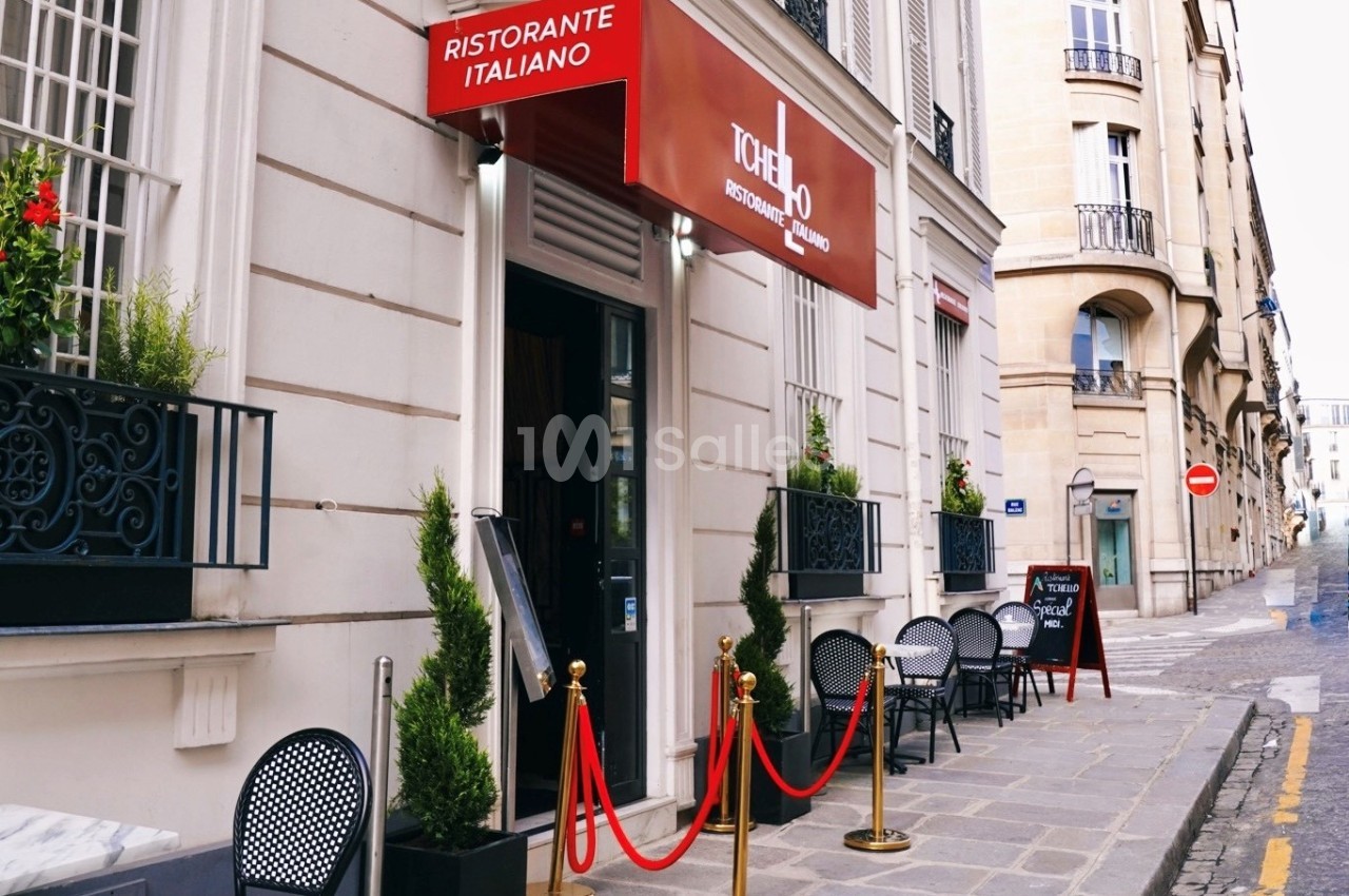 Façade d'un restaurant italien avec terrasse, enseigne rouge et plantes en pots, situé dans une rue pavée.