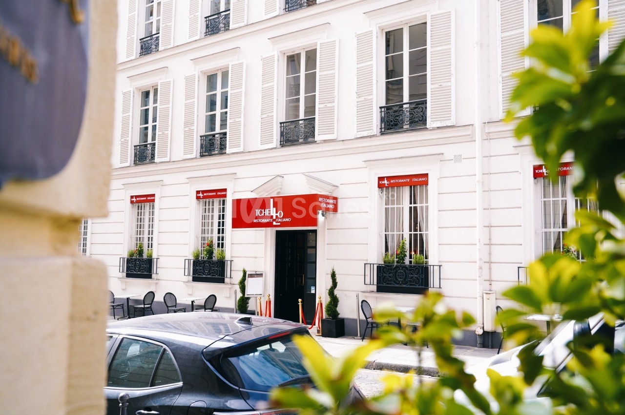 Façade d'un restaurant avec terrasse, stores rouges et fenêtres à volets blancs, vue partielle à travers des feuillages.