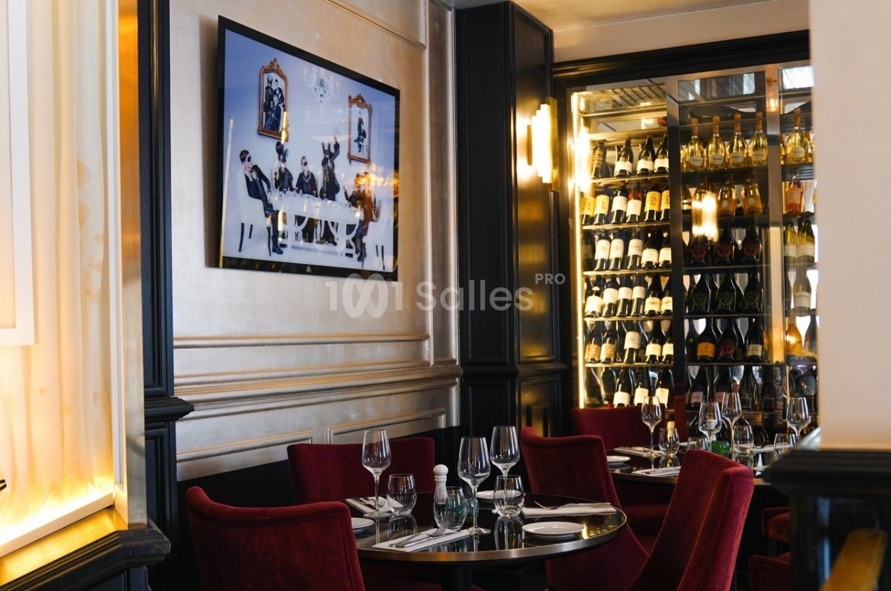 Salle de restaurant élégante avec tables dressées, fauteuils rouges, tableau mural et cave à vin vitrée en arrière-plan.