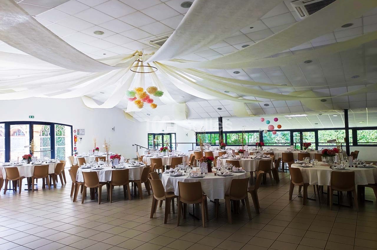 Salle de réception décorée avec des tables rondes dressées, des chaises en bois et des guirlandes suspendues au plafond.