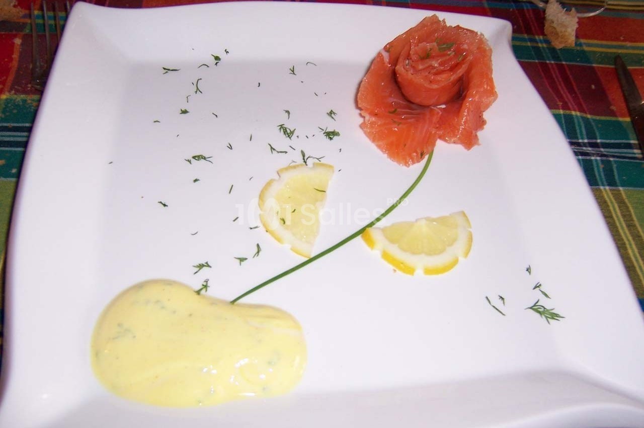 Tranche de saumon fumé disposée en forme de fleur avec citron, aneth et sauce jaune sur une assiette blanche.