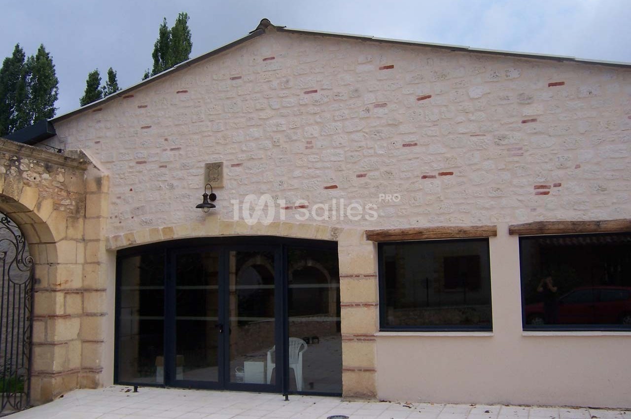 Façade en pierre claire avec grandes fenêtres vitrées et encadrements en bois, sur une terrasse pavée.