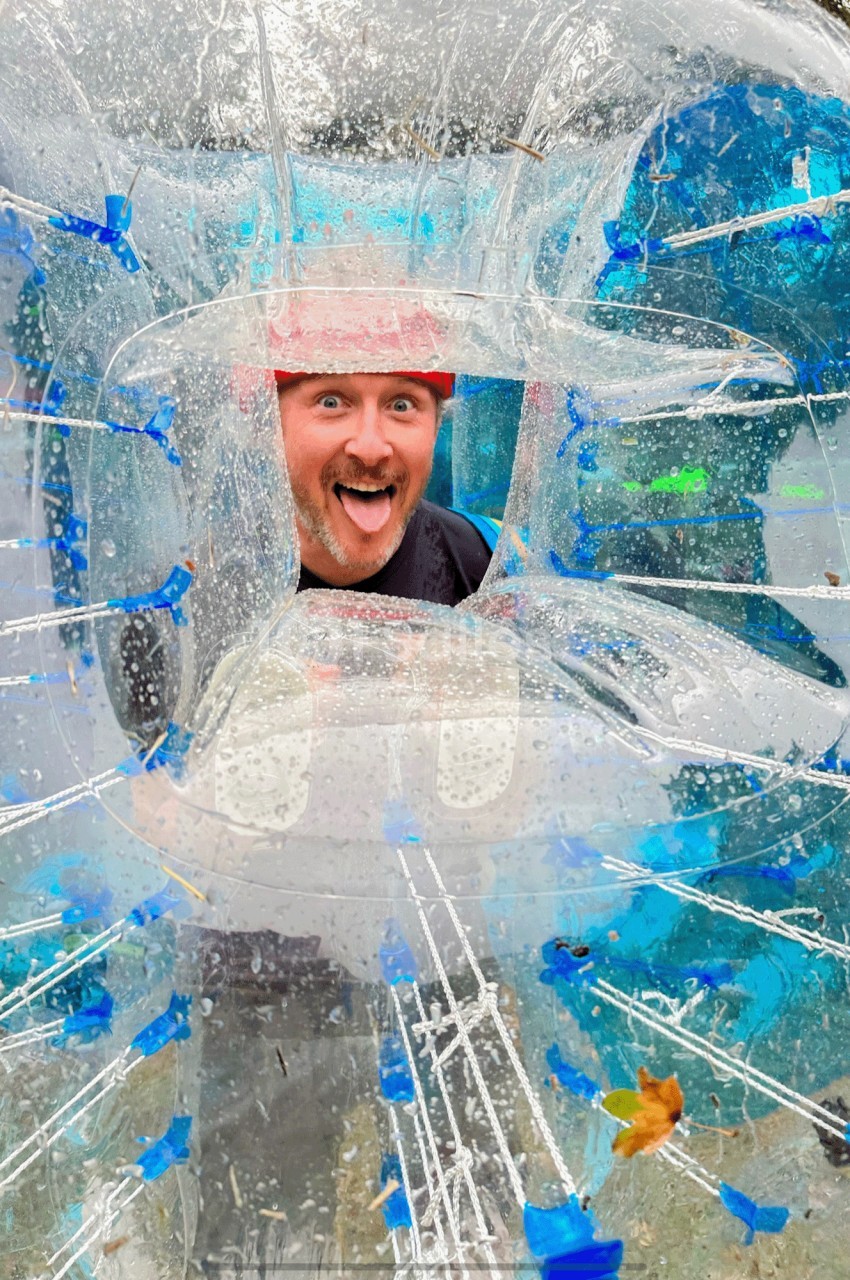 Homme souriant à travers une bulle gonflable transparente, portant un casque rouge, dans un cadre extérieur.